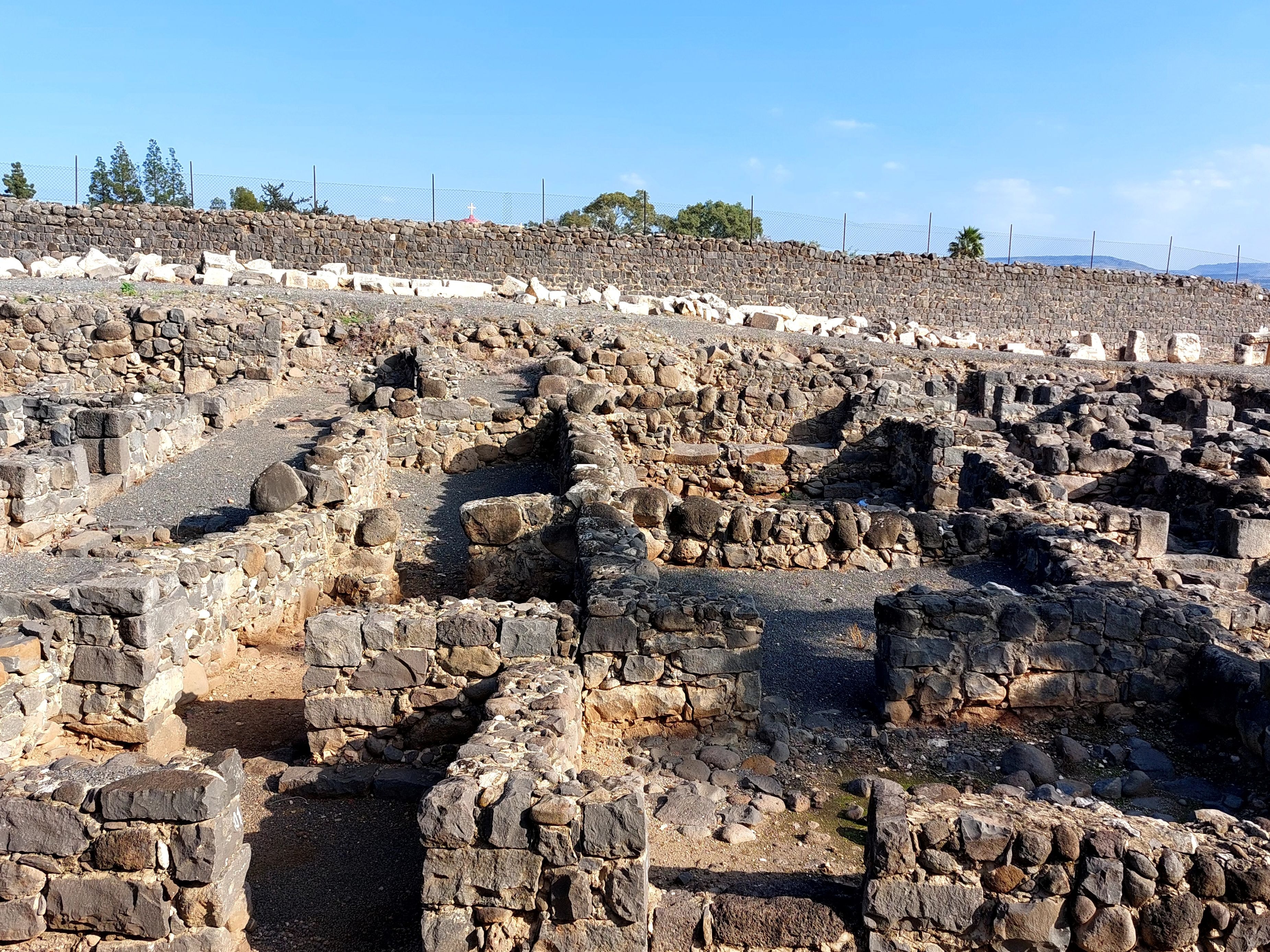 Ancient stone ruins at Capernaum with a long wall in the background running parallel to the Roman road