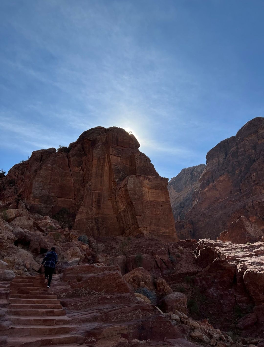 A lone figure climbing ancient stone steps at Petra with the sun glowing behind towering rose-red sandstone cliffs