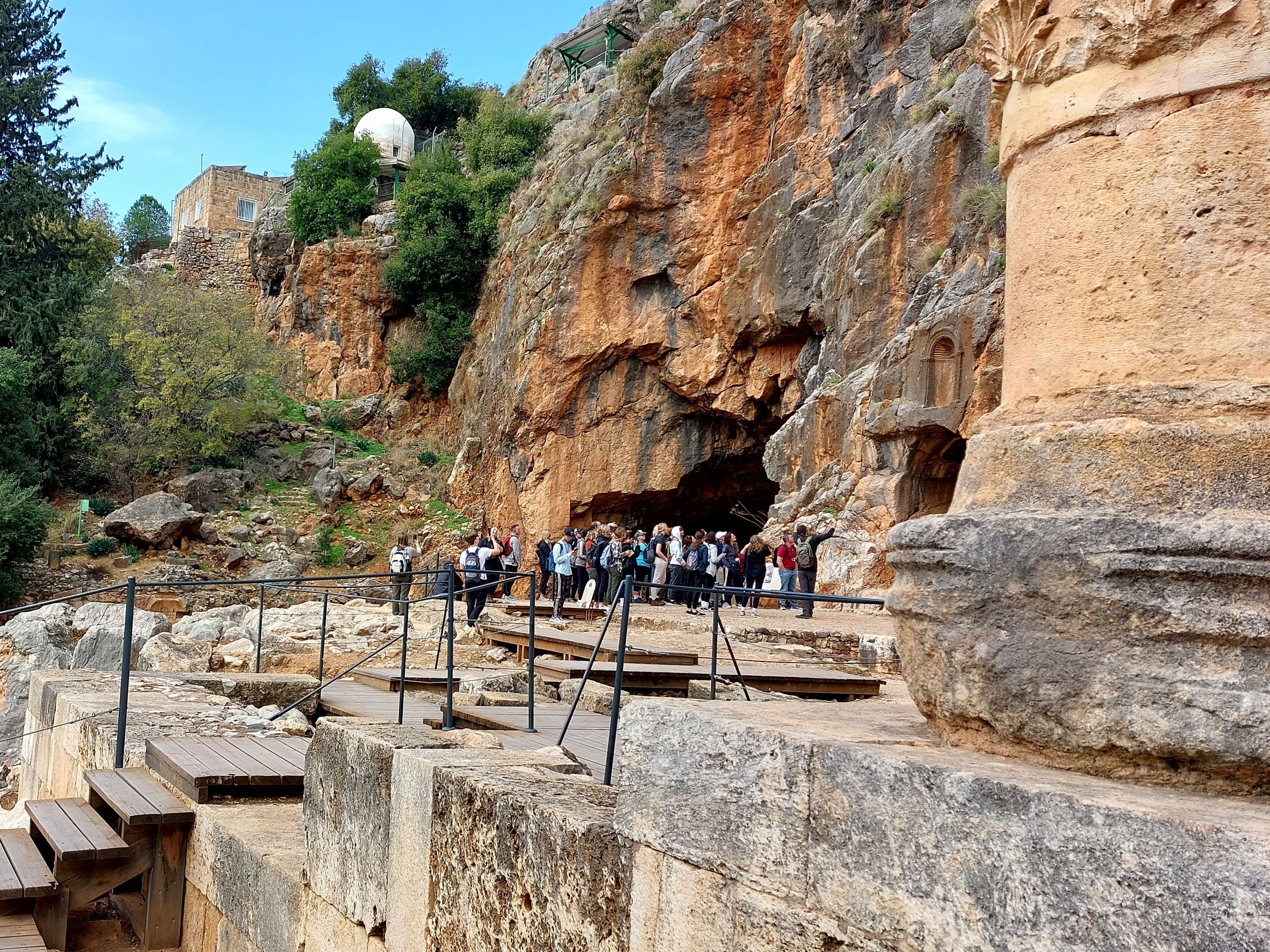 Tour group gathered at the entrance of the cave of Pan at Caesarea Philippi, with carved Roman temple niches visible in the dramatic cliff face beside the cave opening