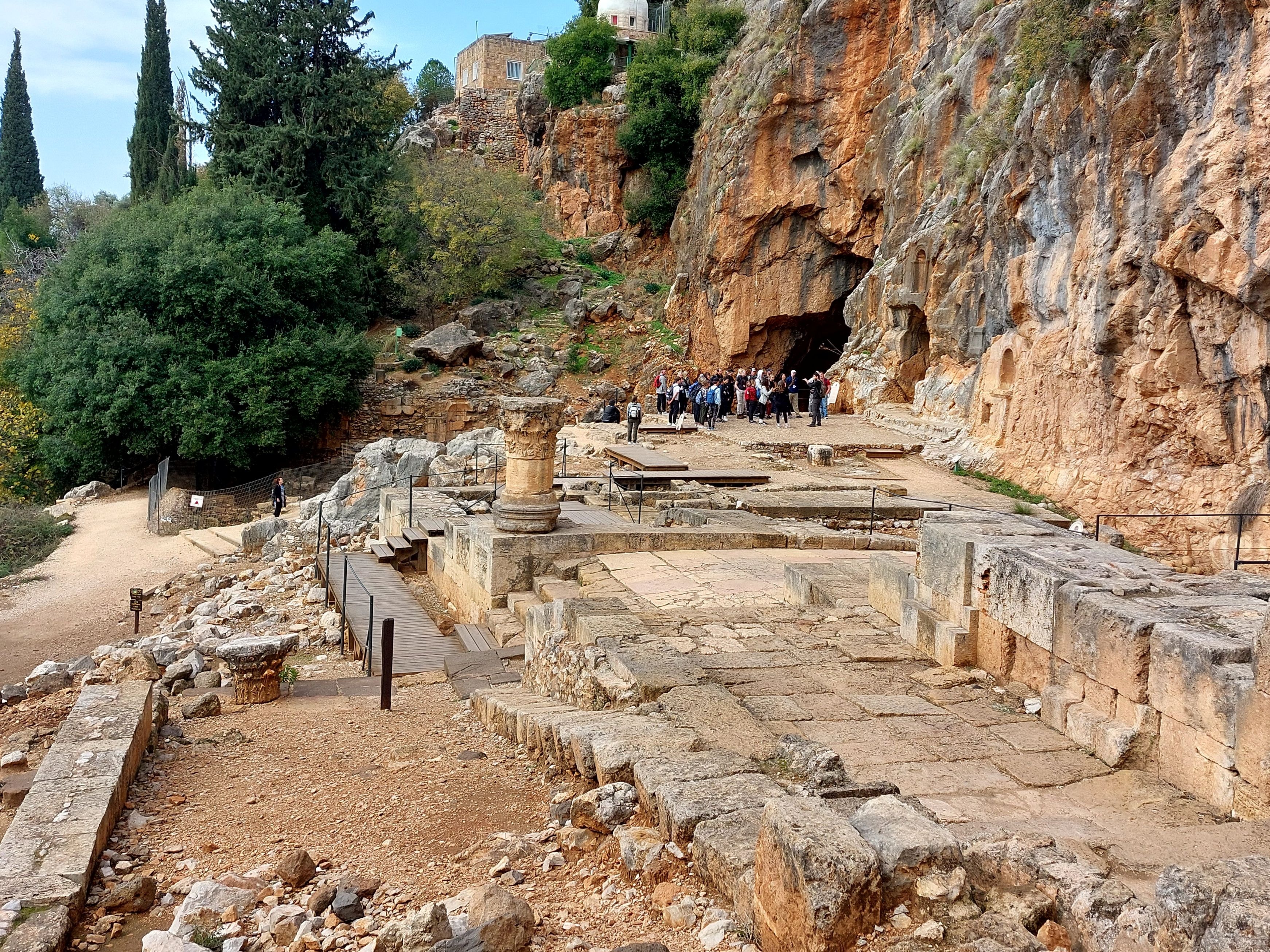 Ruins of the Roman temples at Caesarea Philippi with stone foundations and a lone column standing before the cave of Pan carved into the dramatic cliff face, visitors exploring the site