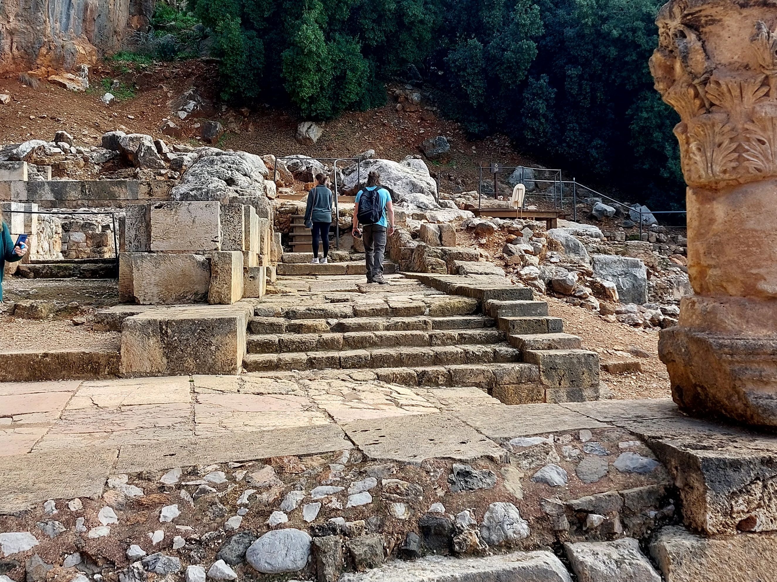 Visitors climbing ancient stone steps through the ruins at Caesarea Philippi with the dramatic cliff face and lush trees in the background, a carved column capital visible in the foreground