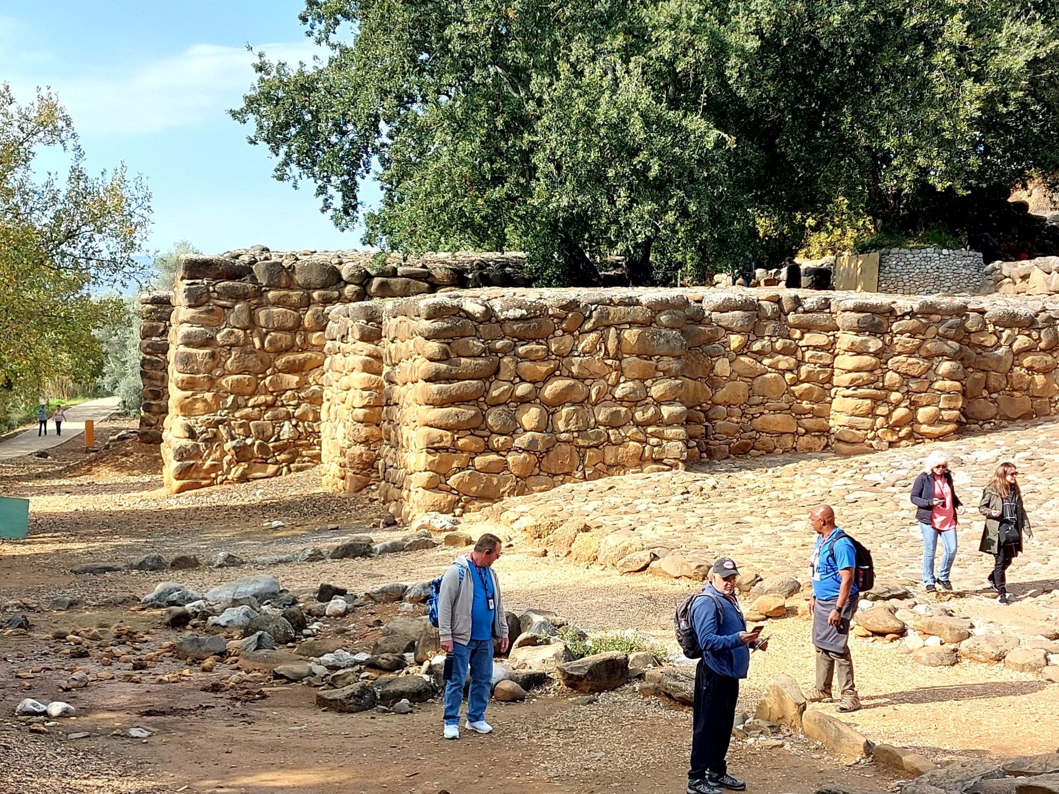 Massive ashlar stone fortress walls of the ancient city of Dan from the time of Jeroboam, with tour group members standing at the base examining the well-preserved stonework beneath large trees