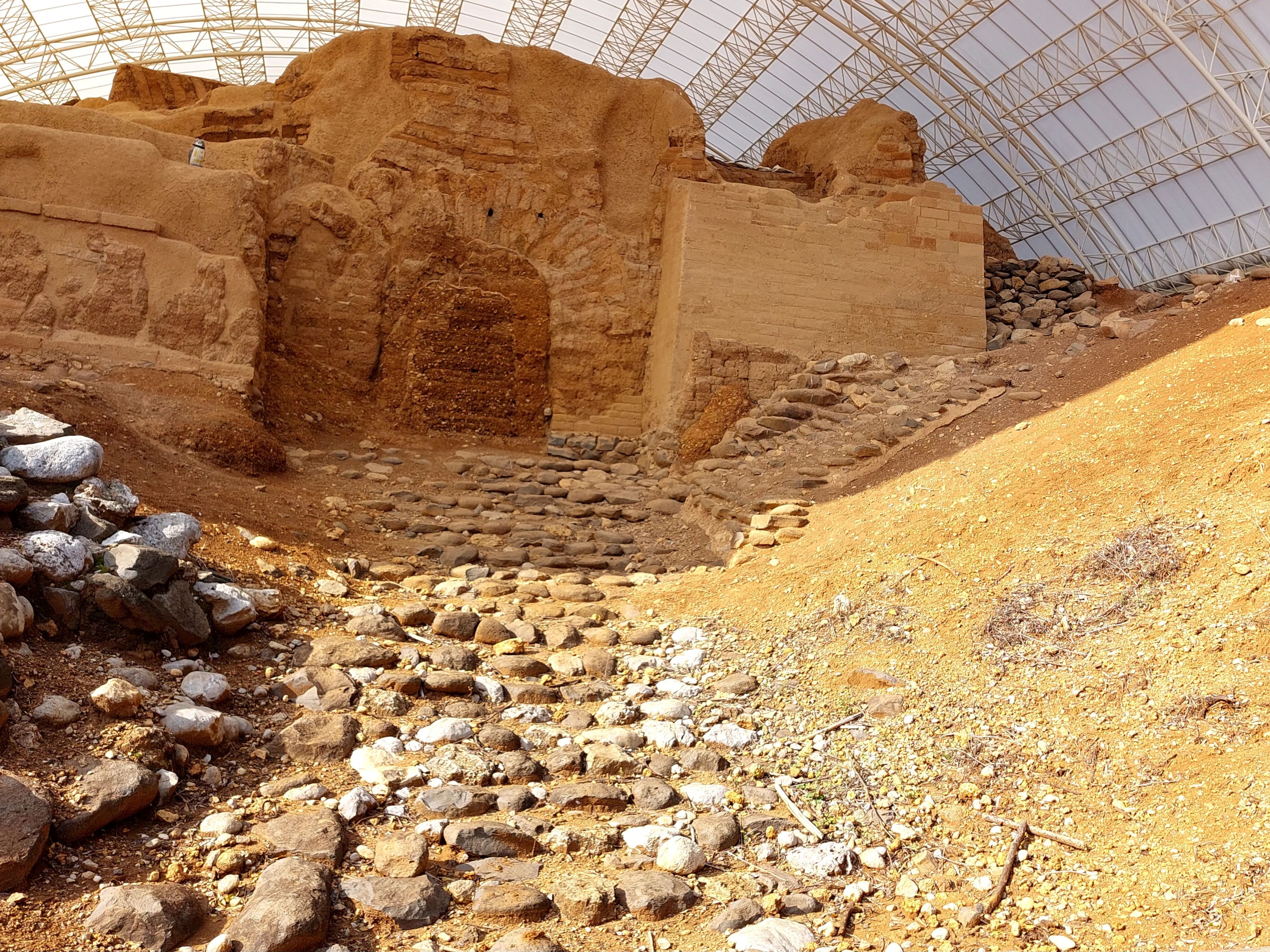 Ancient mud-brick gate structure at the city of Dan preserved under a protective dome, with a cobblestone ramp leading up to the sealed entrance and earthen mounds on either side
