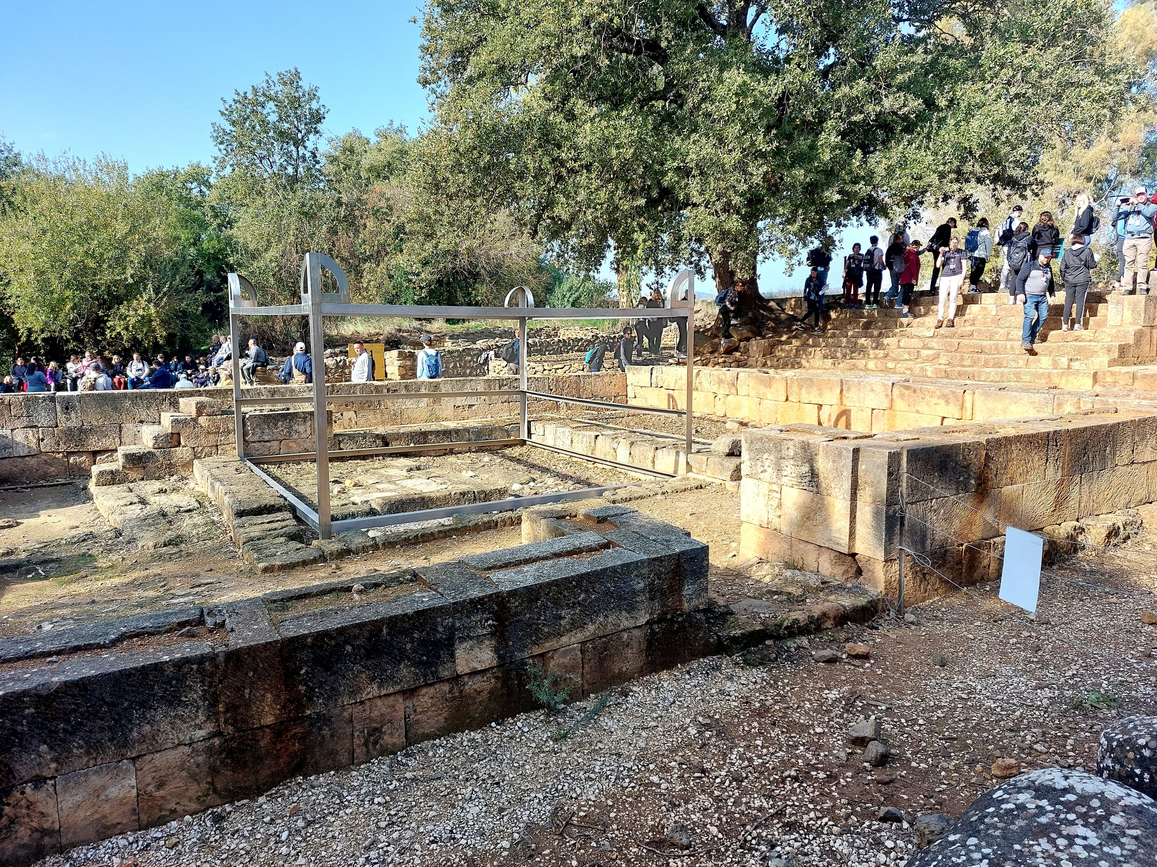 Ruins of the ancient city of Dan showing the stone platform of Jeroboam's altar with a modern metal framework outline marking its original dimensions, surrounded by tour groups on the ancient stone steps beneath large trees