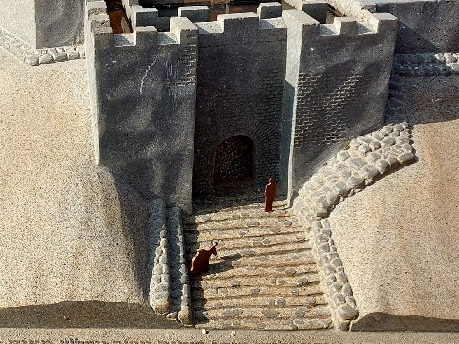 Detailed architectural model of the ancient city of Dan showing the arched city gate, cobblestone ramp, stone walls with battlements, and tiny figures of a person and a donkey entering the city