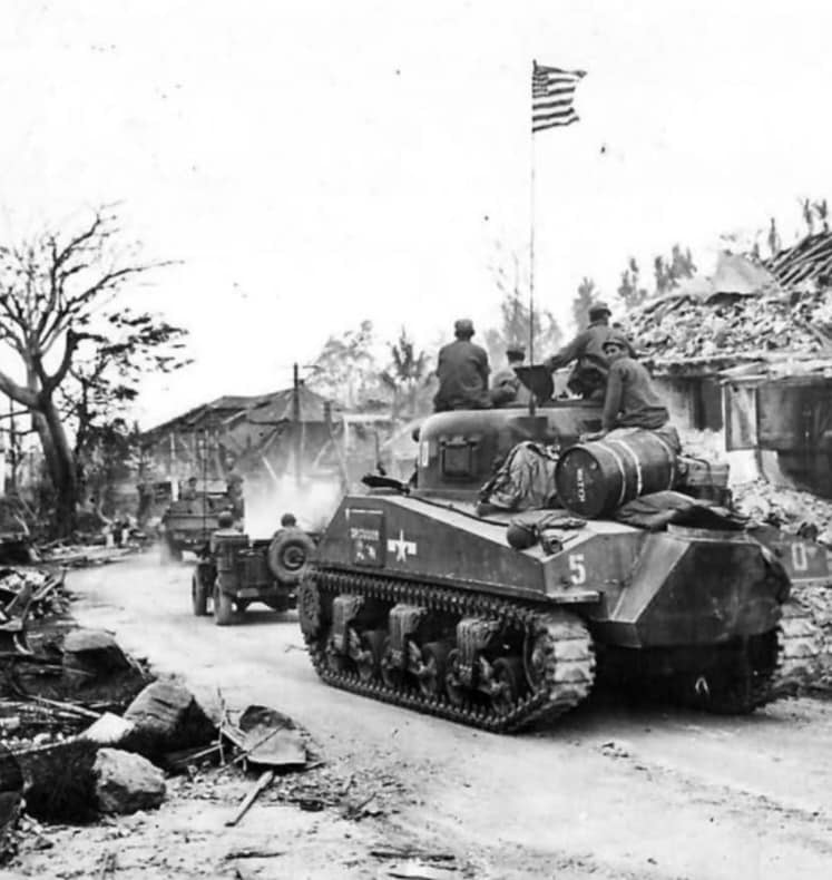 American tank rolling through a destroyed French town with the American flag flying above during Operation Nordwind